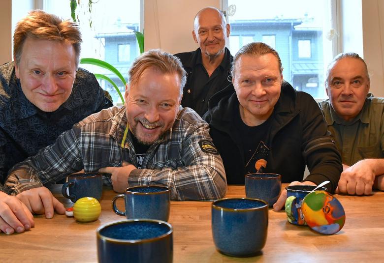 Kuvassa ovat yhtyeen viisi jäsentä vaalean ruskean pöydän ääressä istumassa eri värisissä paidoissa. Edessä on sinisiä mukeja.