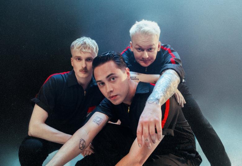 Kuvassa ovat Kuumaa-yhtyeen kolme jäsentä istumassa tiiviisti yhdessä. Heillä on tummat t-paidat päällään. Tausta on tumman sininen.