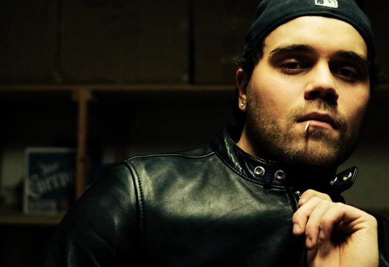 Kuvassa on oikeassa reunassa puolivartalokuvassa ja hänestä näkyy mustalla pohjalla kasvot ja vasenta kämmentä. Hänellä on musta nahkatakki päällään.