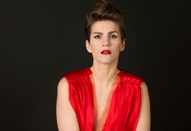 Kuvassa on Maria Ylipää istumassa puolivartalokuvassa. Hänellä on punainen, hihaton mekko päällään ja siinä on v-kaula-aukko. Tausta on tumma.