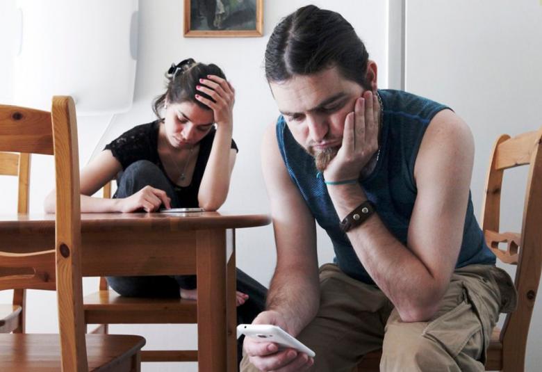 Kuvassa on etualalla oikealla nuori mies istumassa, nojaa vasemmalla kädellään poskeensa ja katsoo oikeassa kädessään olevaa kännykkää. Pöydän takana istuu nuori nainen ja katsoo myös kännykkäänsä. 