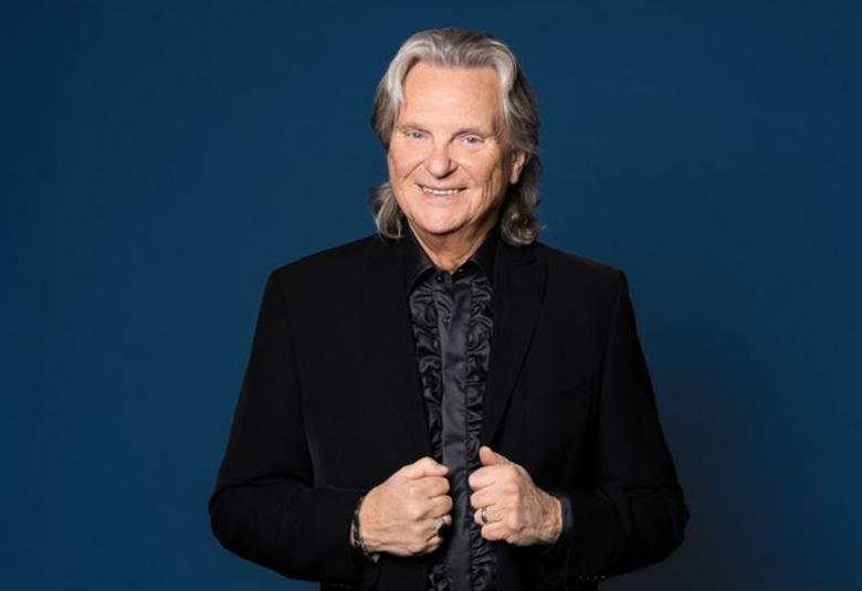 Kuvassa on keskellä Pepe Willberg ja hänellä on musta takki sekä musta paita. Hän pitää molemmilla ksillän kiinni takin liepeistä. Tausta on sininen. 