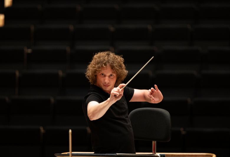 Kuvassa on Santtu-Matias Rouvali johtamassa orkesteria tahtipuikko silmiensä tasalla puolivartalokuvassa. Hänellä on musta t-paita päällään. Tausta on musta.