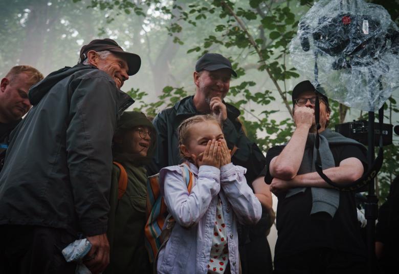 Kuvassa on keskellä vaalean sinisessä farkkutakissa Siiri Virtanen ja hänen ympärillään on kuvausryhmän kuusi henkilöä tummissa ulkovaatteissa. Yläpuolella on vihreitä lehtipuun oksia ja kaikki seisovat puun alla.