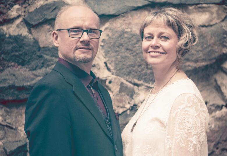 Kuvassa on vasemmalla Tapio Lappalainen  mustassa takissa ja Sanna Lappalainen vaaleassa puserossa. Taustalla on kiviseinää, jossa on suuria, tummia kiviä ja vaaleita välimuurauksia. 