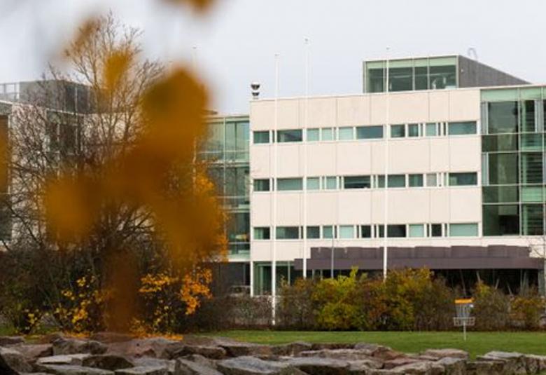  Kuvassa on vasemmalla olevan lähes lehdettomän puun takana oleva rakennus, joka on L.n muotoinen ja jonka oikea osa on vaalea. Alla näkyy litteähköjä kiviä ja pihapensaita