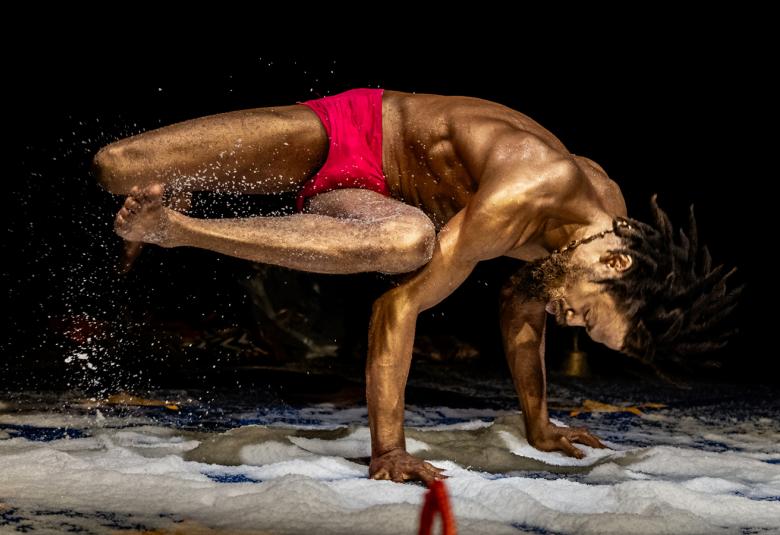 Kuvassa mies, jolla on pronssiseksi maalattu iho, ja hän käsien päällä ja muu keho vinottain vaaka-asennossa ylhäällä. Hänellä on punaiset alushousut. Tausta on musta ja lattialla on valkoista vaahtoa.