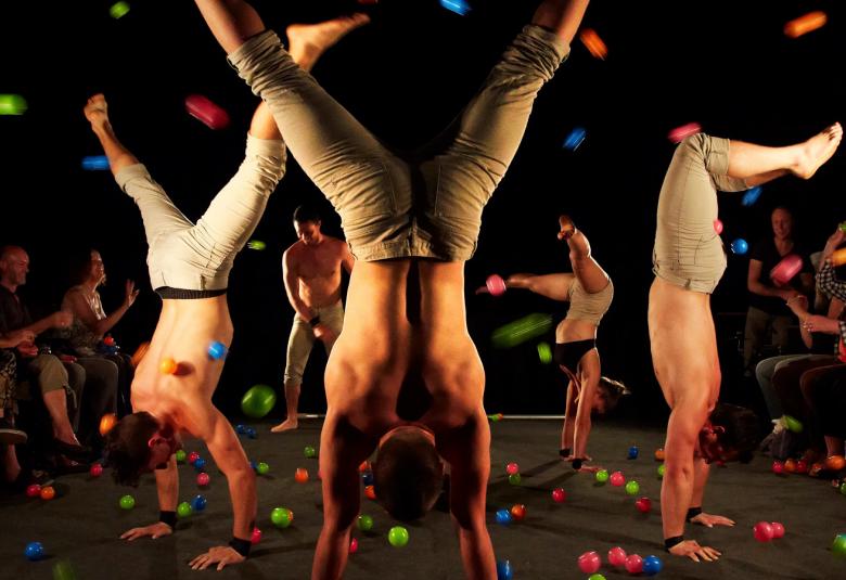 Kuvassa on näyttämöllä kolme miestä ja yksi nainen seisomassa käsillään ja heillä on vaalean harmaat pitkät housut päällään. Yksi mies seisoo taustalla. Esiintyjät ovat lavalla ja yleisö on samalla tasolla. Pimeässä tilassa on eri värisiä valoja. 