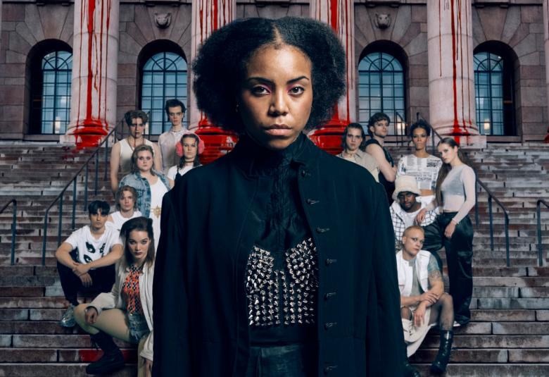 Kuvassa on keskellä mustassa mekossa Abdirahman Bile iso kokoisena ja hänen takanaan on joukko muita näyttelijäitä ryhmänä rapuilla punertavan tiilirakennuksen edessä. 