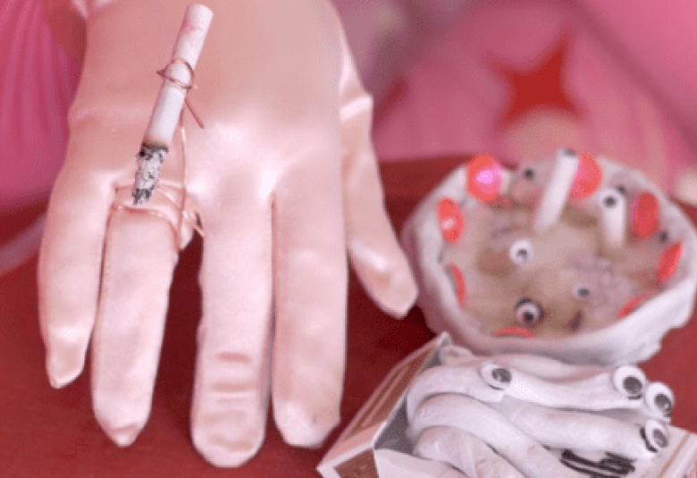 Kuvassa on pinkillä silkkisellä pohjalla käsi vaaleassa hansikkaassa ja sen päälle on kiinnitetty tupakka. Oikealla on epämääräisen muotoinen valkoinen tuhkakuppi, jossa on vaaleita keraamisia tumppeja. Edessä käyristettyjä keraamisia tupakoita.