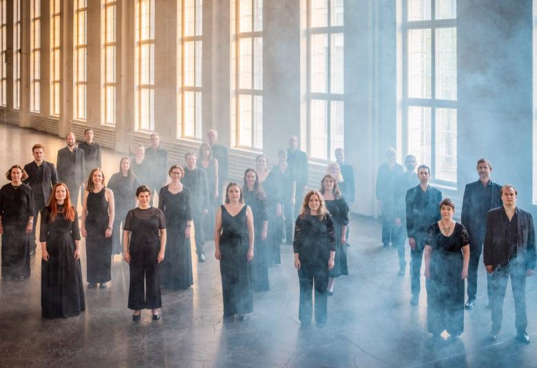 Kuvassa on Helsingin kamarikuoron jäsenet seisomassa mustissa mekoissa ja puvuissa. He ovat isossa hallissa, jossa on korkeat ruutuikkunat rivissä, joista tulee valoa.  