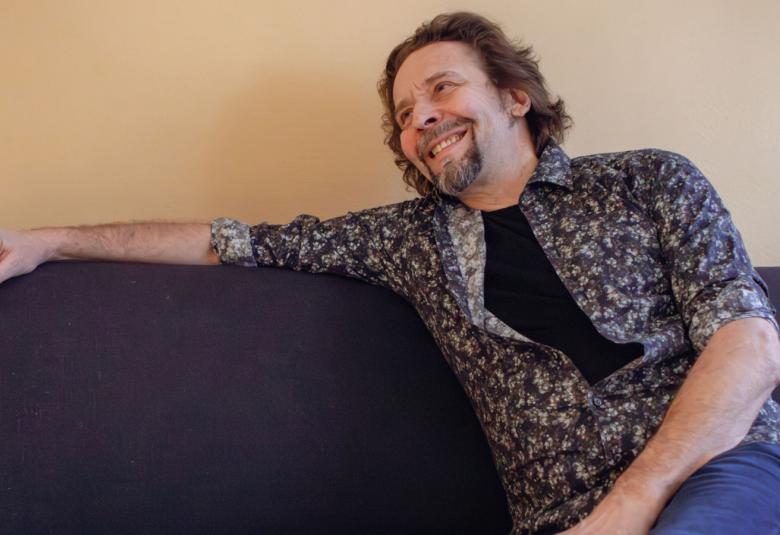 Kuvassa on Ilari Johansson istumassa mustalla sohvalla ja hänen oikea kätensä on ojentuneena sohvan selkänojan päällä. Hänellä on mustan kirjava paita päällään. Seinä on kellertävä.