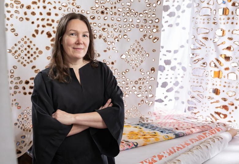 Kuvassa on vasemmalla Johanna Pöykkö mustassa mekossa puolivartalokuvassa. Taustalla on ripustettu kankaita, joissa on ruskeareunaisia reikiä erilaisina kuvioina. 