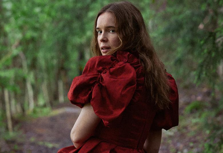 Kuvassa on Marketta Tikkanen selin kävelemässä metsätietä, jonka varrella on koivuja. Tkkasella on punainen mekko päällään ja siinä on puhvihihat.