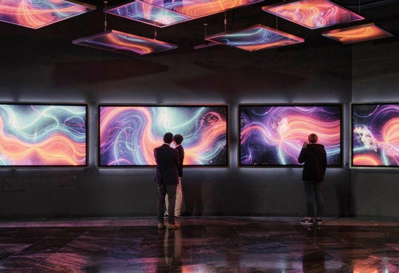 Kuvassa on keskellä ruuturivissä värikkäitä kiemurtelevia viivoja ja pintoja.  Tila on tumma ja kuvat heijastuvat kattoon ja lattiaan. Kuvia katselee pariskunta ja yksittäinen katselija.