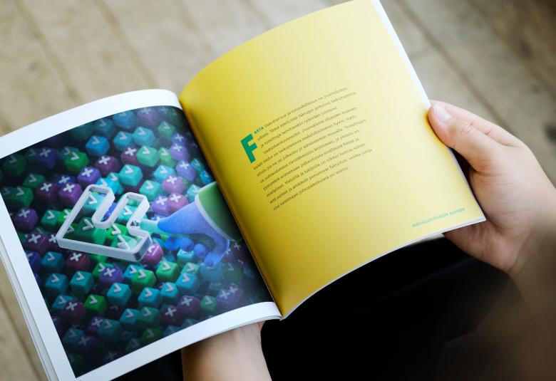 Kuvassa on kädet pitelemässä kirjaa, jossa vasen sivu on tummansininen ja siinä on kirjaintunnuksia. Oikealla keltaisella sivulla on keskellä puoli sivua tekstiä.