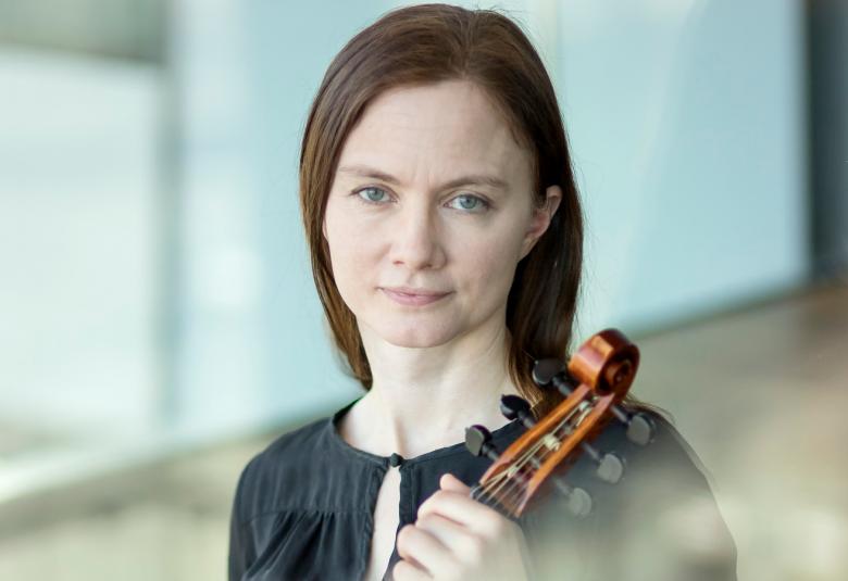 Kuvassa on Maija Lampela puolivartalokuvassa ja hännellä on viulu kädessään. Hän on pukeutunut murtaan  puseroon. Taustalla on vaalean sinistä seinää.