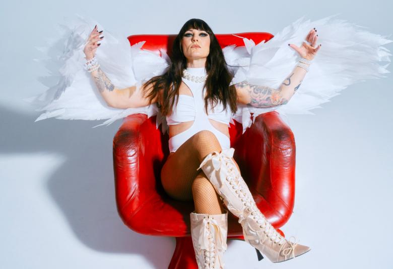 Kuvassa on Mira Luoti istumassa punaisessa kiiltävässä nojatuolissa. Hänellä on valkoinen mekko ja kädet levitettyinä sivuille ja niissä on enkelin siivet. Tausta on vaalea ja siinä on varjo vasemmalla.