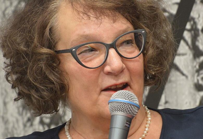 Kuvassa on Monika Fagerholm kasvokuvassa ja puhumassa mikrofoniin. Hänellä on ruskeat kiharaiset hiukset ja silmälasit.