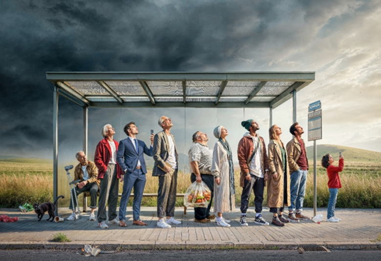 Kuvassa on ihmisiä bussipysäkillä, jossa on valokate.  Kaikki katsovat taivaalle, jossa on tummaa pilveä ja vaalenvaa taivasta oikealla. Taustalla on keltaista viljapeltoa.