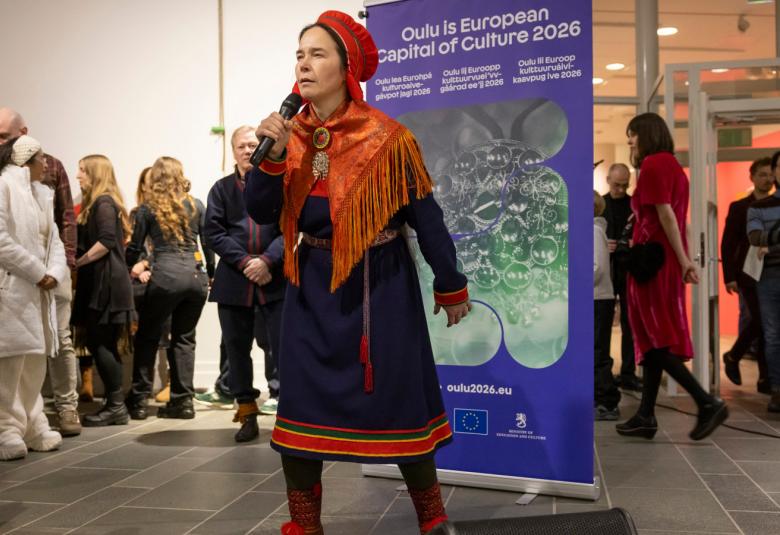 Kuvassa on Anna Morottaja keskellä kuvaa esiintymässä Lapin puvussa, joka on tumman sininen ja siihen kuuluu puna-oranssi -kuvioinen huivi ja päähine.  Taustalla näkyy Oulu2026 roll-up ja vasemmalla yleisöä.