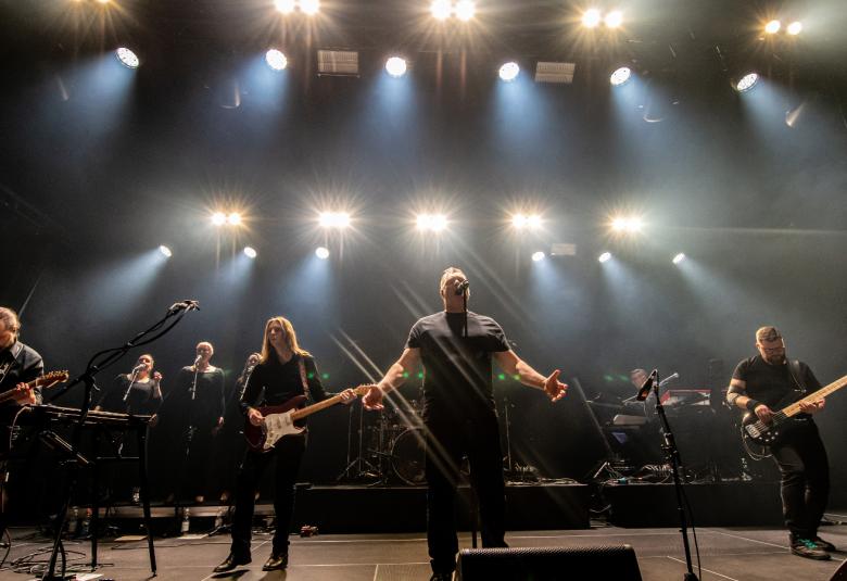 Kuvassa on neljä bändin jäsentä ja kaksi taustalaulajaa.  Lava on tumma ja taustalla näkyy valorivejä. 