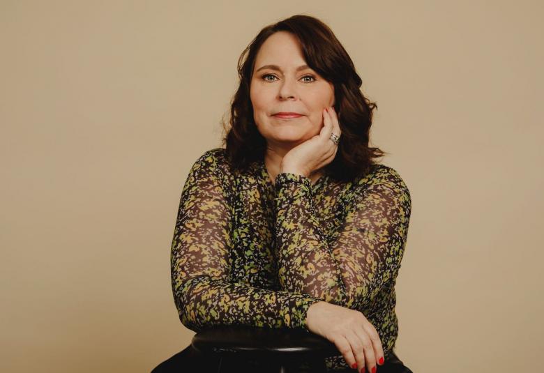 Kuvassa on Saila Susiluoto ja hän istuu sekä nojaa tuolin selkämykseen. Hänellä on ruskean mustan kirjava puseoro päällään.  Kuvan tausta on ruskehtavan harmaa. 