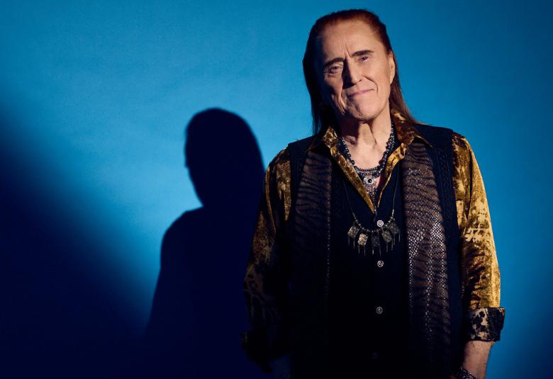 Kuvassa on Tommi Läntinen puolivartalokuvassa seisomassa. Hänellä on ruskea pusero ja musta liivi päällään. Taustalla näkyy hänen varjonsa. Vasemmalla ja alhaalla on tummennosta ja muu osa taustaa on sininen.