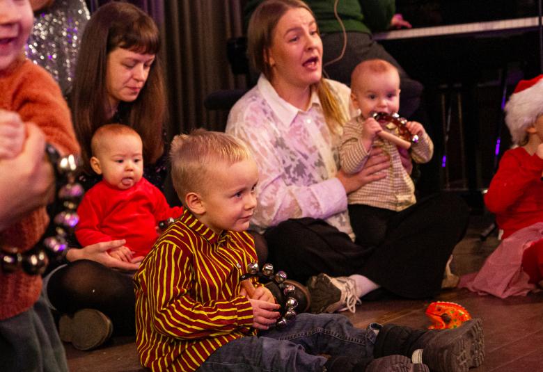 Kuvassa on äitijä ja vauvoja sekä pieniä lapsia istumassa lattialla.  He katsovat esitystä.