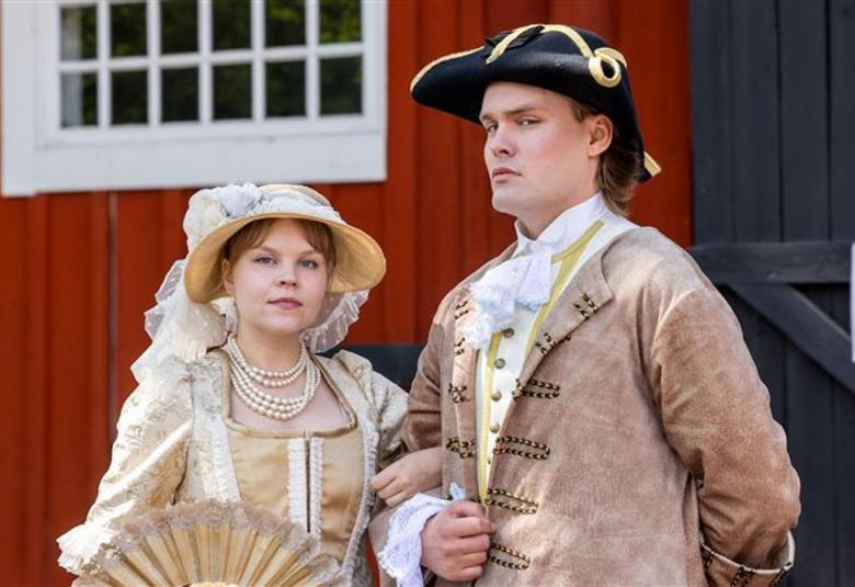 Kuvassa on vasemmalla nuori nainen vaalean ruskeassa vanhanaikaisessa mekossa ja lierihatussa. Asussa ja hatussa on valkoisia röyhelöitä ja brodyyrejä. Oikealla on nuori mies, jolla on musta aatelisen suikkahattu päässään sekä koristeellinen vaalean ruskea takki päällään ja valkoinen röyhelöpaita. Taustalla on punaisen puurakennuksen seinää ja valkoinen, pieniruutuinen ikkuna.