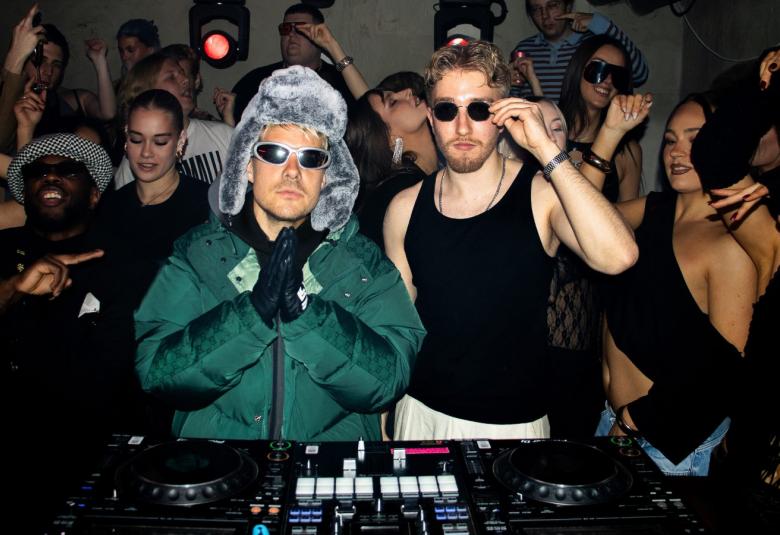 Kuvassa on keskellä vasemmalla VilleGalle vihreässä toppatakissa ja harmaassa karvalakissa sekä aurinkolaseissa. Oikealla hänen vieressään on mustassa hihattomassa t-paidassa sekä aurinkolaseissa. Taukana on nuorta yleisöä. Edessä on musiikin ohjauspöytä. 