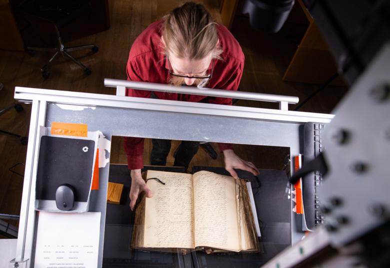 Kuvassa on ylhäältä päin kuvattuna punaisessa puserossa oleva mies, joka pitelee kirjaa alustalla. Päällä on harmaa metallikehikko, jolla digitoidaan kirjaa.