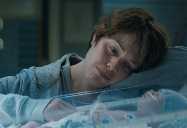 Kuvassa on Oona Airola, joka katselee keskoskaapissa nukkuvaa vauvaa. Hänellä on vaalean sininen pusero päällään. 