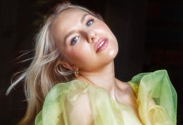 Kuvassa on Ilta puolivartalokuvassa ja hänellä on pää taivutettuna vasemmalle.  Vaaleat hiukset lentävät vasemmalle. Hänellä on päällään vaalean vihreä sifonkinen mekko.