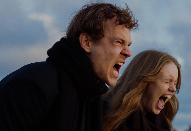 Kuvassa ovat nuori mies ja nainen puolivartalokuvassa ulkotakeissa. He molemmat ovat sivuttain ja huutavat ulkona. Taustalla on puolipilvistä taivasta.