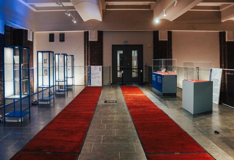 Kuvassa on Helsingin päärautatieaseman näyttelytila, jossa on isossa tilassa molempien seinien luona lasiset vitriinihyllyt. Seinät ja lattia ovat vaalean ruskeat ja keskellä on kaksi pitkää punaista mattoa.