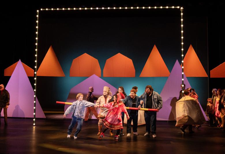 Kuvassa on tummalla näyttämöllä takana punaisia kartiomaisia telttoja. alemana on näytteljöitä tanssimassa edessä värikkäissä vaatteissa ja taaempana seisoo harmassa puvuissa olevia näyttelijöitä. Vasemmalla on kartiomainen lila teltta.