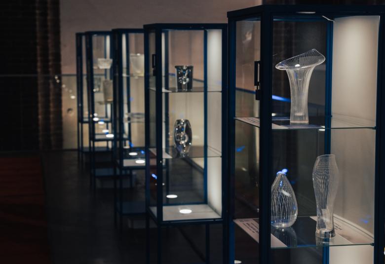 Kuvassa on tummassa tilassa keskellä läpinäkyvästä lasista hyllykkö, jossa on Tapio Virkkalan lasitöitä.