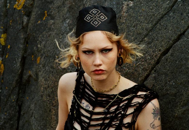 Kuvassa on Vilma Jää puolivartalokuvassa. Hänellä musta, hihaton löyhä verkkopaita päällään ja musta päähine päässään. Taustalla on tummaa kalliota.