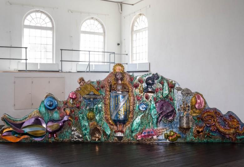 Kuvassa on isossa valkoisessa tilassa, jossa on vasemmalla ylhäällä isoja, yläosasta pyöreitä ja pikkuryytuisia ikkunoita. Tummalla lattialla on kiiltävävärisiä, erityisesti turkoosia, ruskeaa ja lilaa ison krokotiilimaisen hahmon päällä.  Hahmo nousee ylöspäin ja laskeutuu alas.  Päällä on keraamisia kuvioita ja mm. lintuja.