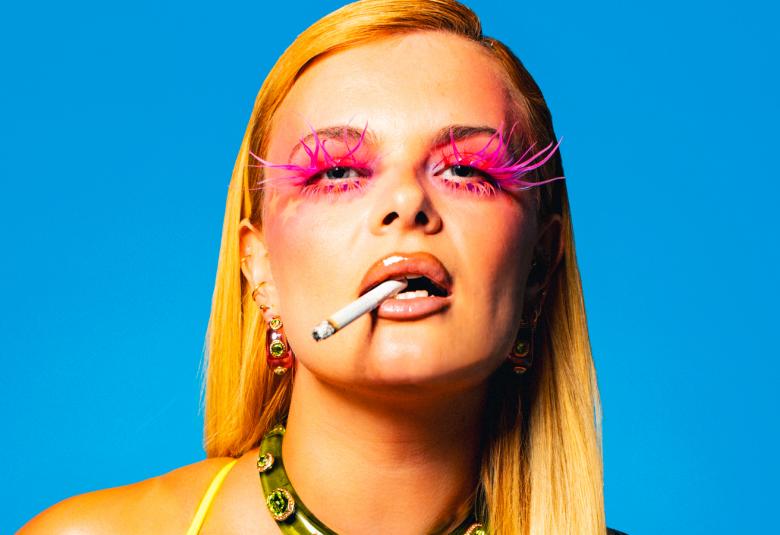 Kuvassa on puolivartalokuvassa Erika Vikman ja hänellä on pitkät vaaleat hiukset, punaiseksi meikatut silmänympärykset ja tupakka hampaissaan. Tausta on turkoosi. 