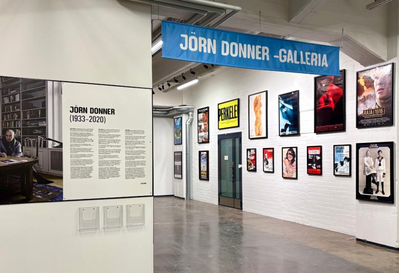 Kuvassa on julistegalleria, joussa on etuseinällä kuvaa ja tekstiosuus. Käytävän toisella puolella on julisteita ja ylläpuolella sininen kyltti, jossa on valkoisin kirjaimin Jörn Donner -galleria.