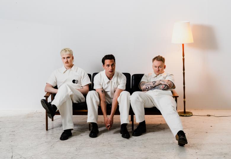 Kuvassa ovat Kuumaa-yhtyeen kolme jäsentä istumassa sihvalla. Heillä kaikilla on valkoiset vaatteet. Oikealla on jalkalamppu. Kuvan tausta on valkoinen ja lattia vaalea.