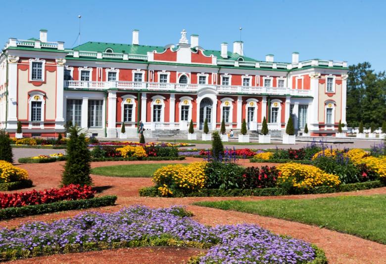 Kuvassa on Kadriorgin taidemuseon rakennus, joka on puna-valkoinen ja siinä on korkeat koristeelliset ikkunat ja valkoinen koko julkisivun yläparveke. Edessä on värikkäitä kukkaistutuksia.