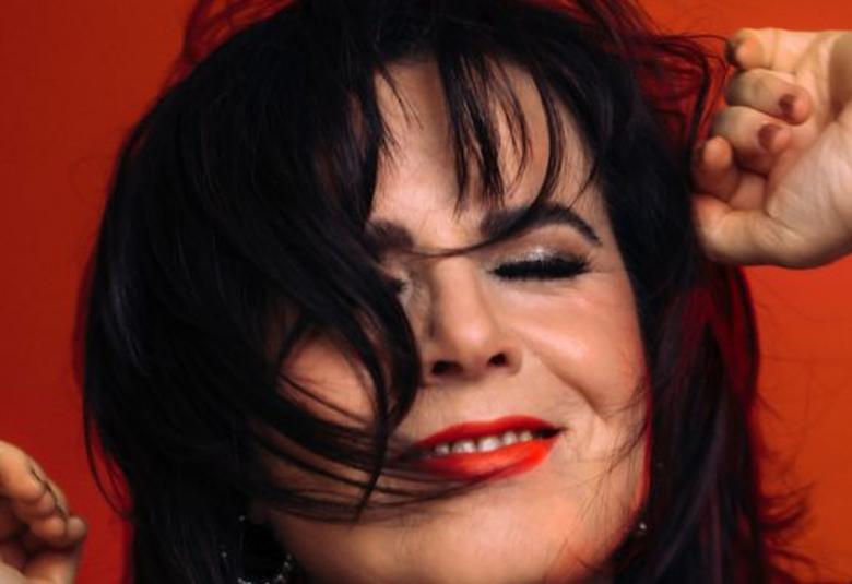 Kuvassa on Kaija Koo kasvokuvassa. Hänellä on tummat hiukset osittain kasvoilla.  Tausta ruskean punainen.