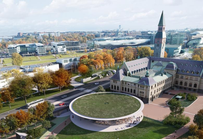 Kuvassa on ilmakuva, jossa on keskellä Kansallismuseon päärakennus ja sen takana pyöreä ruohikkokattoinen, reunasta vaalea rakennus.  Näiden vieressä menee Mannerheimintie ja sen reunassa oransseja, syksyvärisiä puita. Taustalla näkyy kaupunkia.