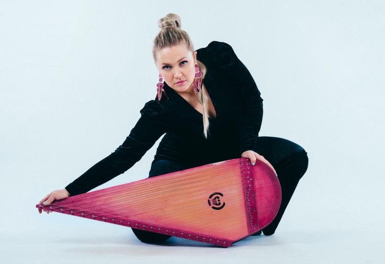 Kuvassa on Maija Kauhanen mustassa asussa kumartuneena punaisen vasemmalle suipoksi muotoillun kanteleen takana ja hän pitää oikealla kädellään kanteleen päästä kiinni. Tausta on vaalean sininen.  