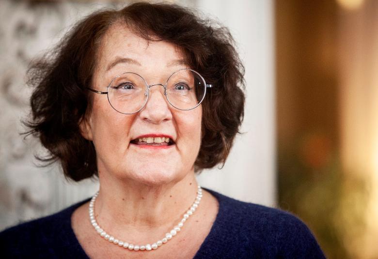 Kuvassa on Monika Fagerholm puolivartalokuvassa ja hänellä on tummat hiukset sekä pyöreät silmälasit. Hänellä on tumma pusero päällään ja valkoiset helmet.