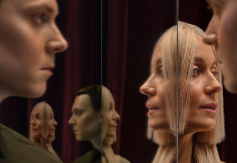 Kuvassa on vasemmalla miehen kasvot sivuttain ja oikealla naisen kasvot sivuttain peilin kautta tuplana samoin taustalla on kasvoja tuplana. Kuvan sävy on tumma.