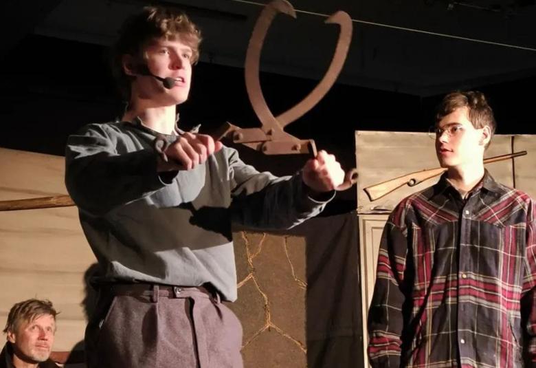 Kuvassa on vasemmalla nuori mies, joka pitää isoja hakasia käsissään ylöspäin.  Vieressä oikealla on toinen nuori mies katsomassa ja taustalla istuu mies.